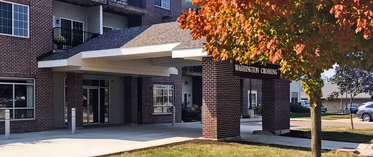 Entrance of a brick building with a covered driveway and a sign that reads 'Washington Crossing'. There is a tree with autumn-colored leaves in the foreground and a few parked cars in the background.