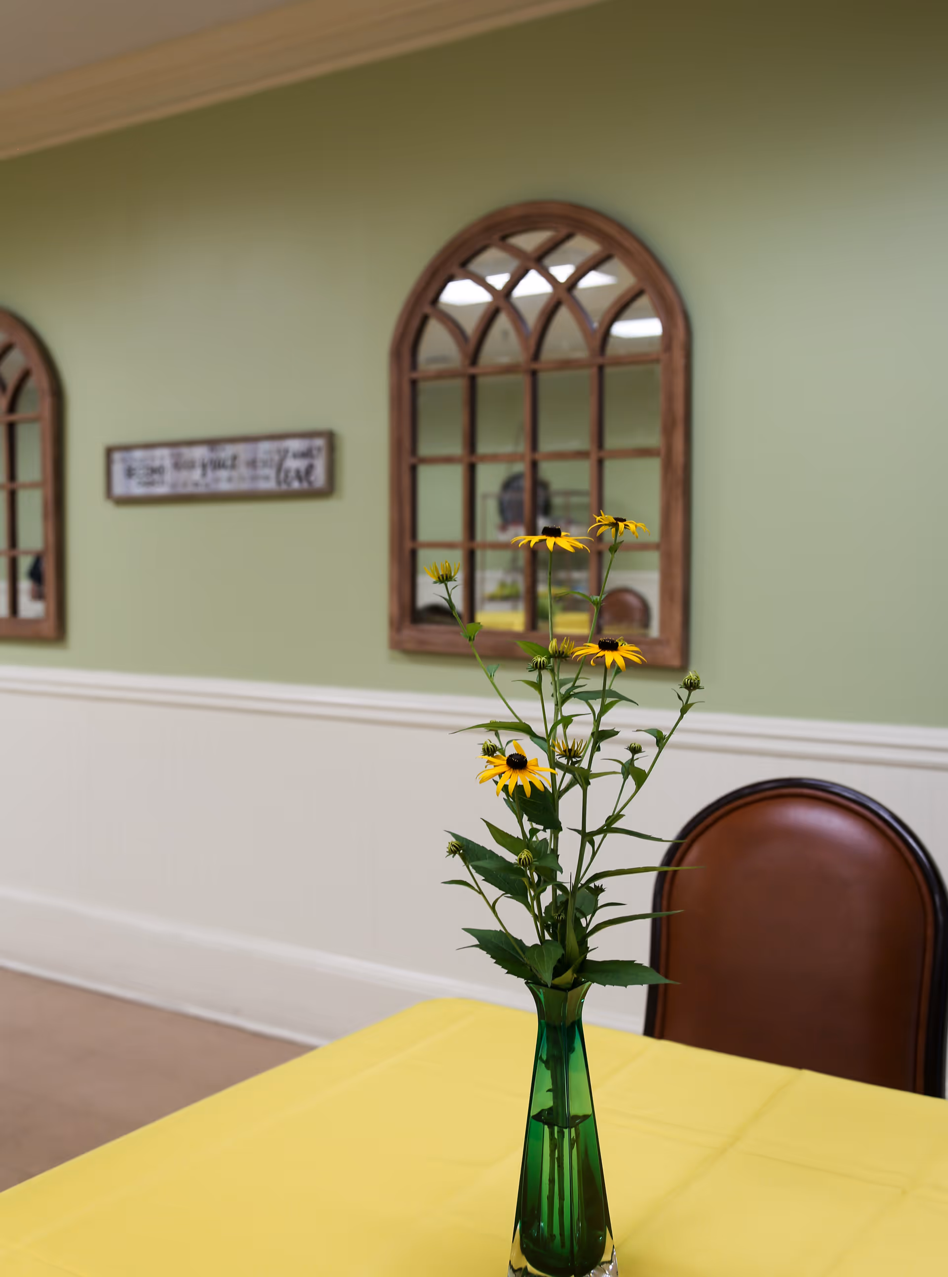 A green glass vase with yellow flowers on a table covered with a yellow tablecloth. Behind the table is a brown chair and a green wall with two arched wooden framed mirrors and a decorative sign.