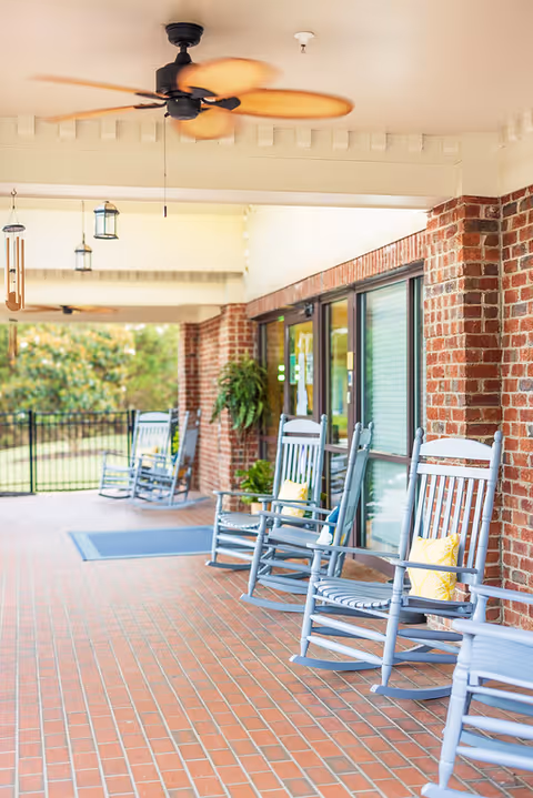 Covered brick porch with several blue rocking chairs, ceiling fans, and a view of a fenced yard.