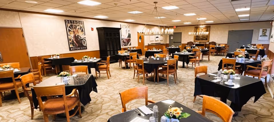 A spacious dining room with multiple tables covered in black tablecloths, each set with cups, glasses, and small floral centerpieces. Wooden chairs surround the tables, and the room features beige walls with vintage Detroit Motor City posters, carpeted floors, and ceiling lights. There is a wooden cabinet against one wall and a chandelier hanging from the ceiling.