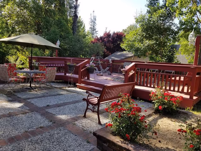 Outdoor patio area with a red wooden deck, a bench, a table with floral cushioned chairs under a large umbrella, and red flowering plants. Trees and greenery surround the area, creating a peaceful garden setting.