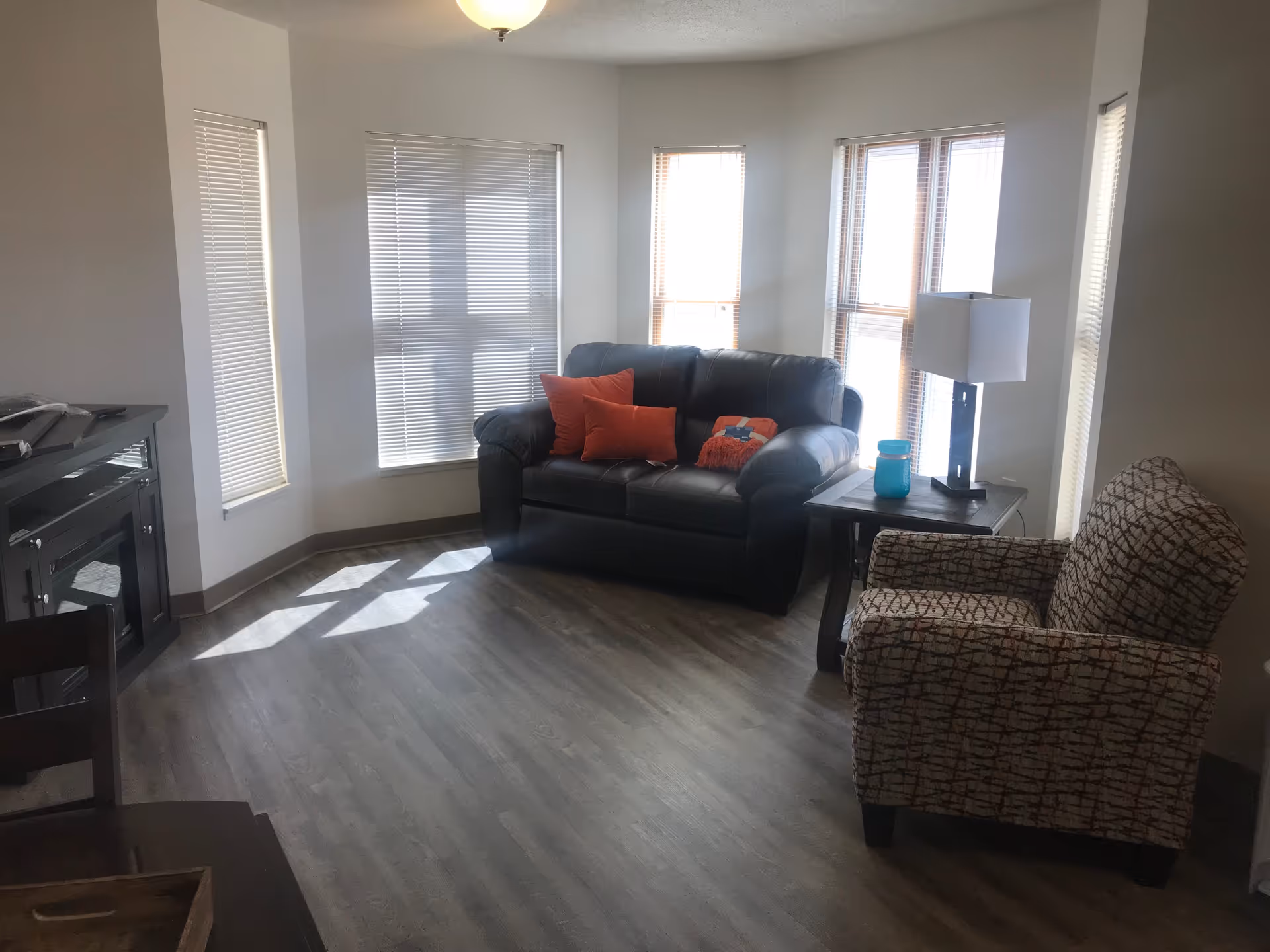 Bright living room with a black leather loveseat, patterned armchair, side table with a lamp, and bay windows with blinds.