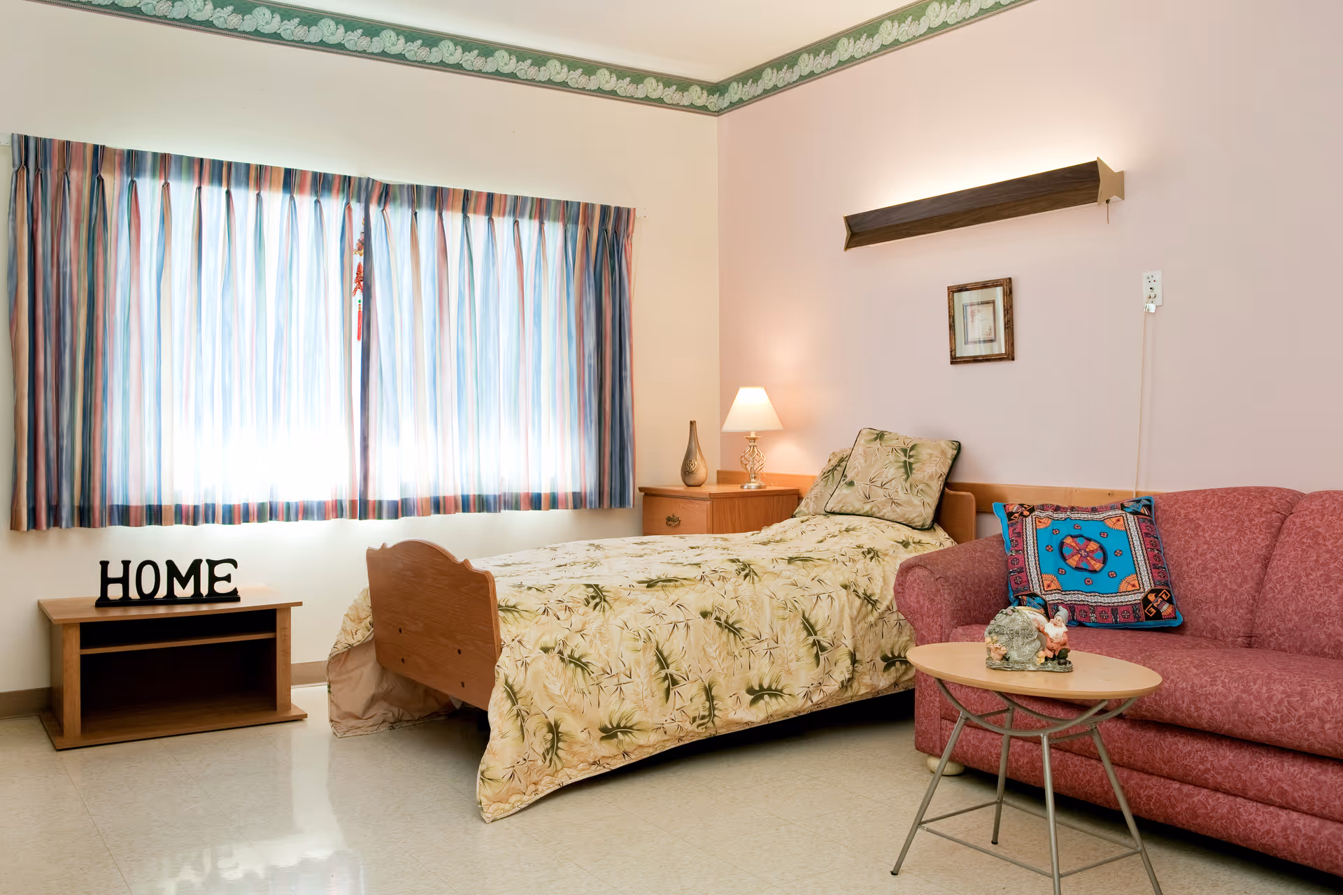 A cozy bedroom in a senior living facility featuring a single bed with floral bedding, a wooden nightstand with a lamp and decorative vase, a small wooden table with the word HOME on it, a pink upholstered couch with a colorful pillow, and a small round table with a decorative item. The room has a large window with striped curtains and light-colored walls with a green patterned border near the ceiling.