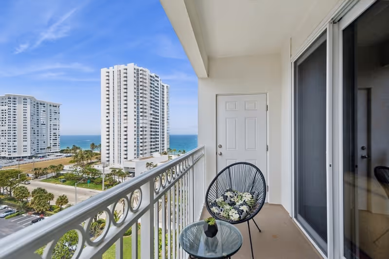 Balcony with a round black chair with floral cushions and a small glass table with a small plant, overlooking a street, palm trees, tall white buildings, and the ocean under a blue sky.