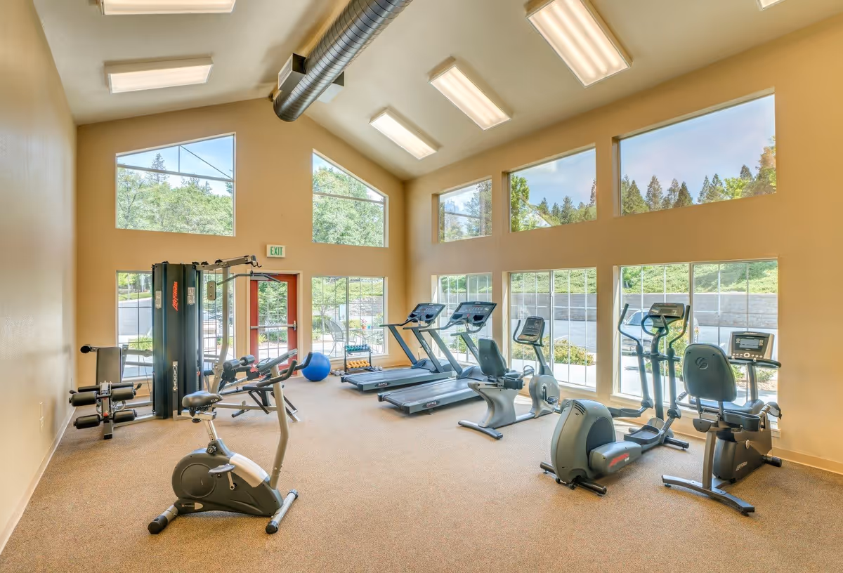 Bright fitness room with large windows showing outdoor greenery. The room contains exercise equipment including stationary bikes, treadmills, elliptical machines, and a multi-function weight machine. The ceiling has exposed ductwork and multiple fluorescent lights.