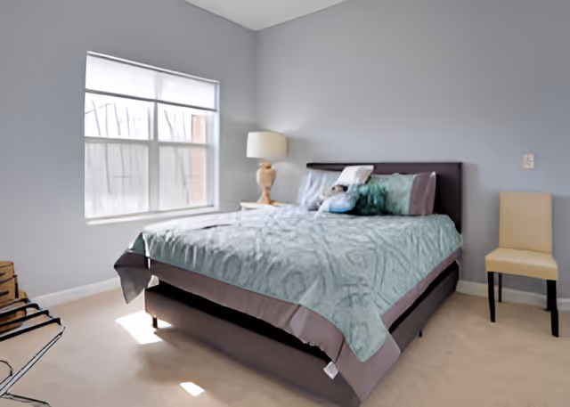 Bedroom with a bed dressed in teal bedding, a bedside lamp, window, and a chair against light blue walls.