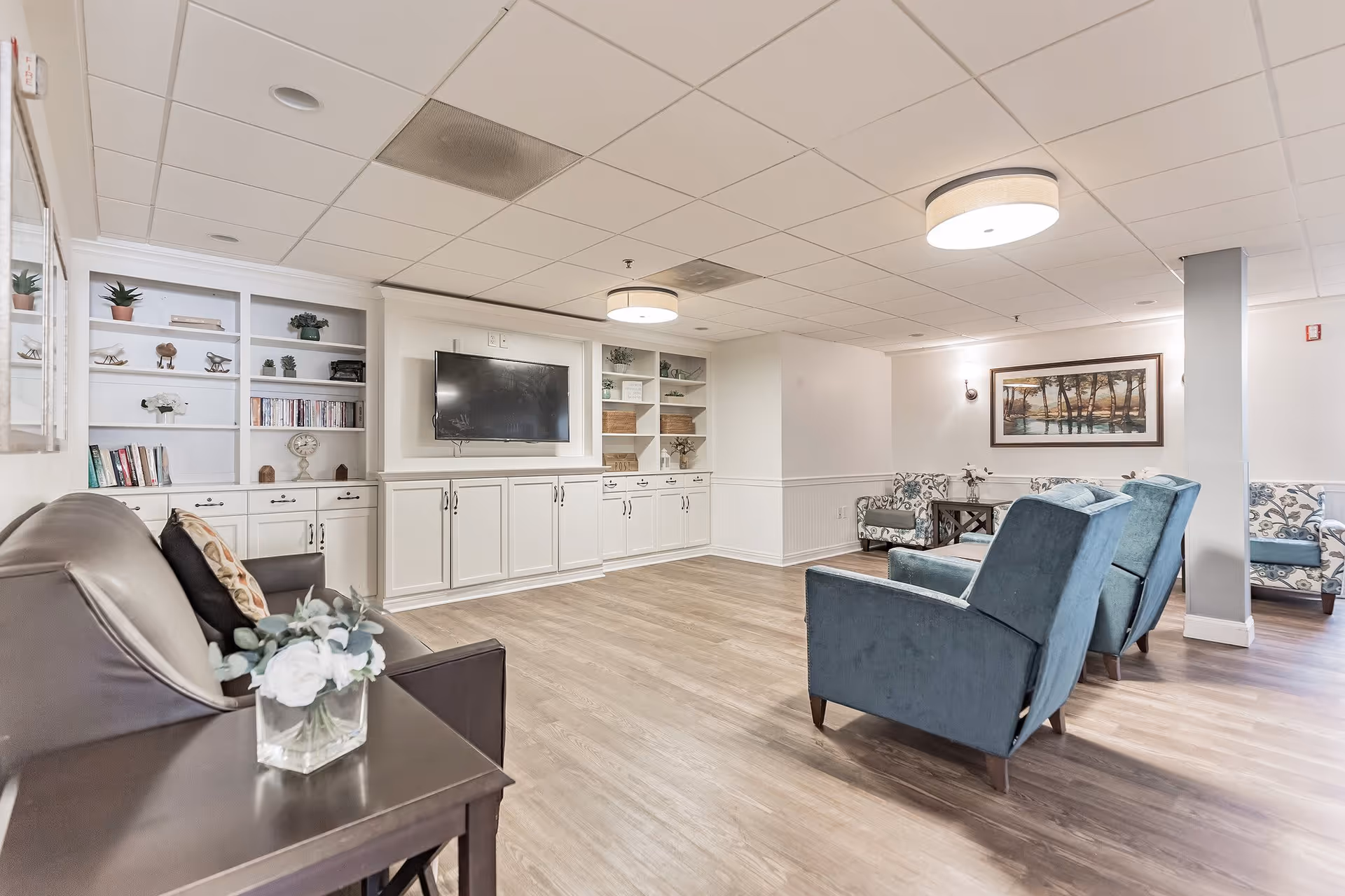 A bright and spacious senior living common area with light wood flooring, white built-in shelves and cabinets, a wall-mounted flat screen TV, and comfortable seating including a brown leather sofa, blue armchairs, and patterned chairs. The room is decorated with plants, books, and artwork, and has modern ceiling lights.