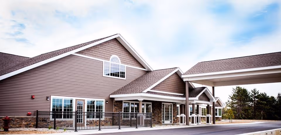 Exterior view of a senior living facility building with brown siding, stone accents, multiple windows, and a covered entrance area. The sky is partly cloudy and there are trees in the background.