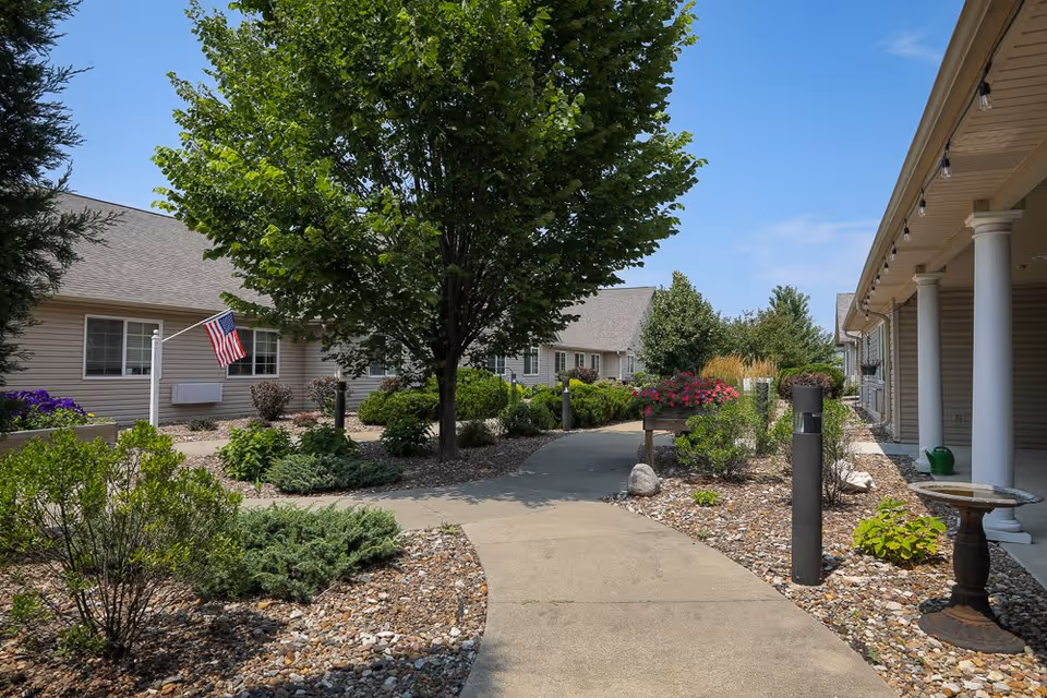 A landscaped outdoor walkway at Homestead Assisted Living & Memory Care of Oskaloosa, featuring a concrete path surrounded by bushes, flowers, and trees. The buildings have beige siding and a gray roof, with an American flag displayed on the left side. There are white columns supporting a covered porch on the right side, and the sky is clear and blue.