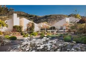Sunlit white modern buildings and patios seen from a landscaped courtyard with rock gardens and hills in the background.