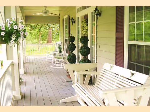 A covered porch with white wooden rocking chairs and a white wooden bench swing. There are potted topiary plants and hanging flower baskets, with a view of green trees and a railing along the porch.