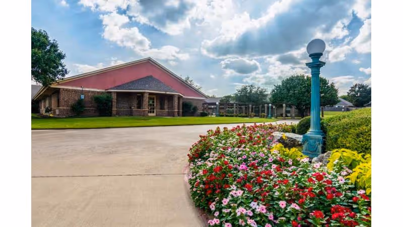 A single-story brick building with a triangular roof under a partly cloudy sky. In the foreground, there is a curved concrete driveway bordered by a colorful flower bed with red, pink, and white flowers, and a decorative green lamp post. Trees and shrubs surround the building and flower bed.