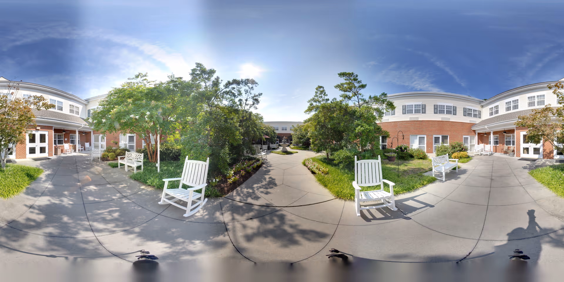 Sunny landscaped courtyard with white rocking chairs, paved walkways, and a two-story brick senior living building surrounding the garden.
