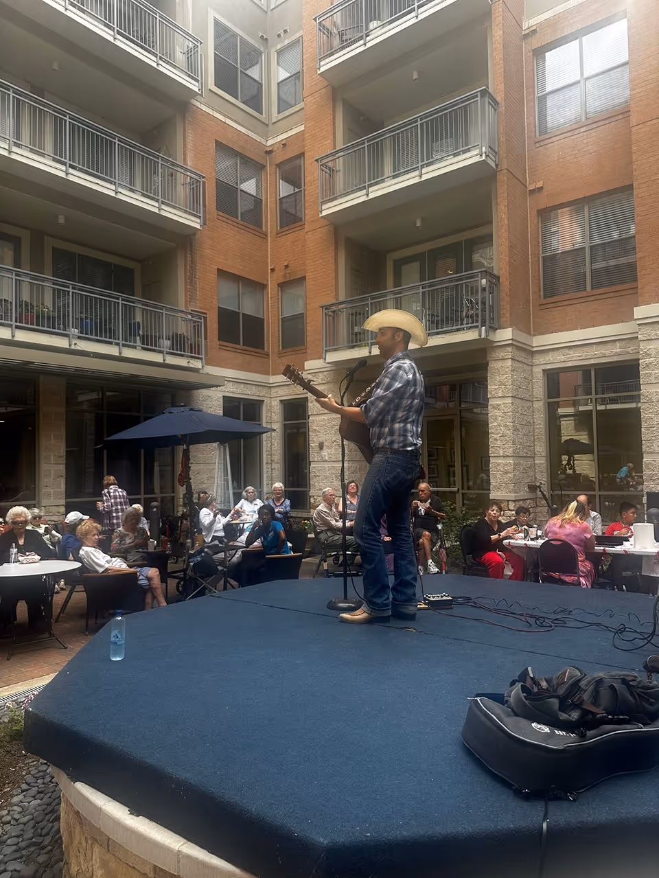 A man wearing a cowboy hat and plaid shirt plays guitar and sings on a small stage in an outdoor courtyard of a senior living facility. Several elderly residents are seated around tables under umbrellas, watching the performance. The courtyard is surrounded by a multi-story brick and stone building with balconies and windows.