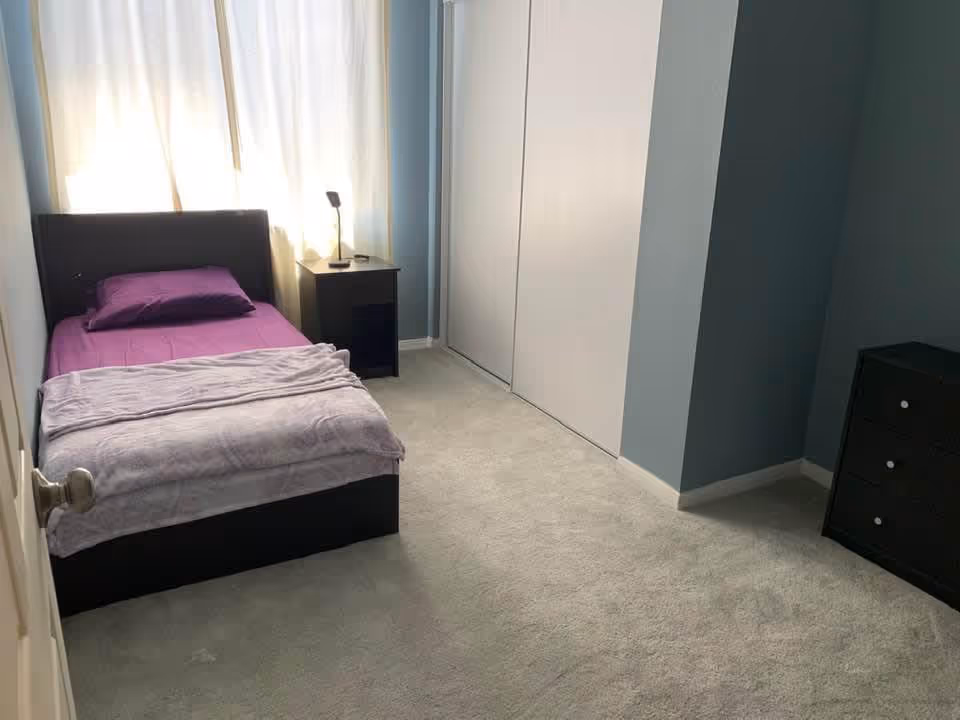 A small bedroom with a single bed made with purple sheets and a light gray blanket. There is a black nightstand with a small lamp next to the bed, a window with light-colored curtains behind the bed, a closet with white sliding doors, and a black dresser with three drawers in the corner. The walls are painted light blue and the floor is carpeted in a light beige color.