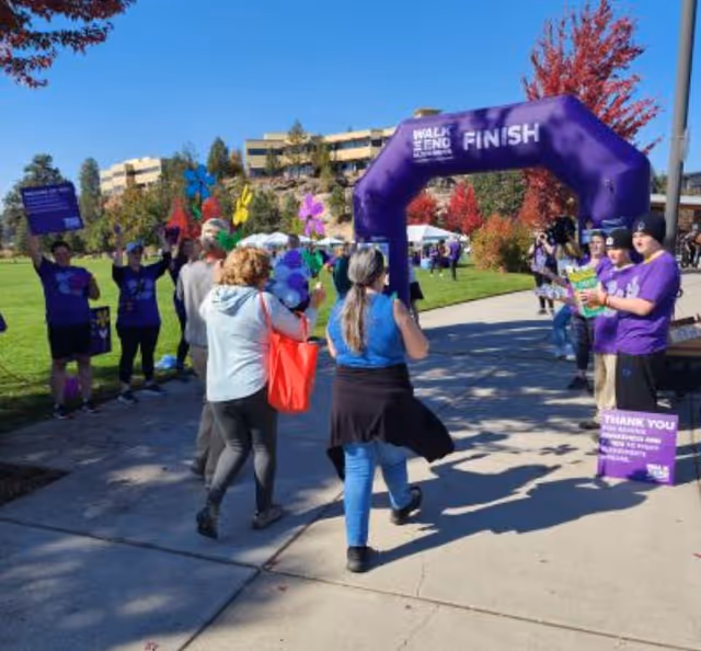 People walking towards a purple inflatable arch that says 'FINISH' at an outdoor event. Several individuals are wearing purple shirts and holding signs and balloons. The event takes place on a sunny day with trees showing autumn colors and buildings in the background.