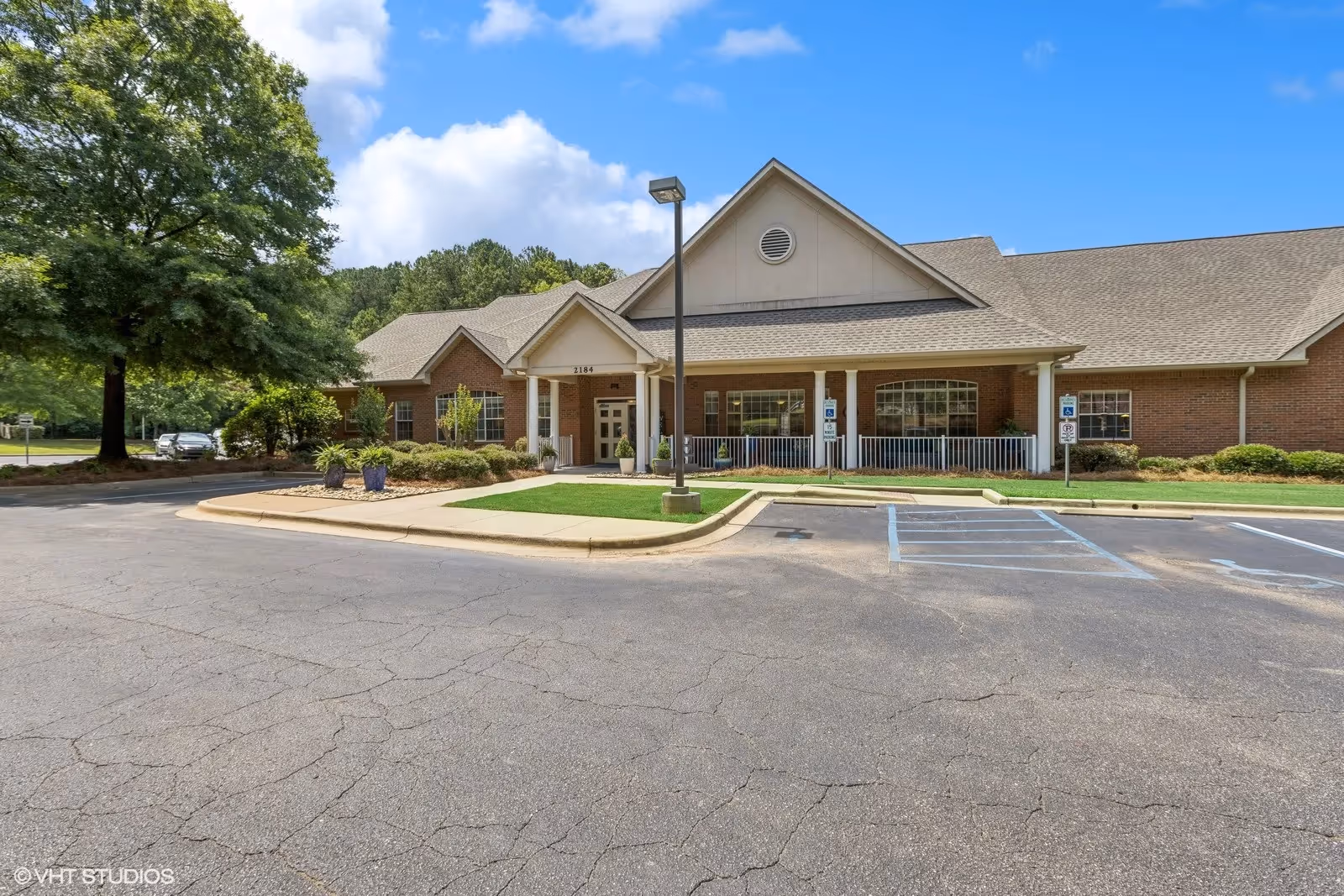 Exterior view of a single-story brick building with a gabled roof and a covered entrance supported by white columns. There is a parking lot with marked handicapped spaces in front, a streetlamp, and some landscaping including trees and shrubs. The sky is clear with a few clouds.