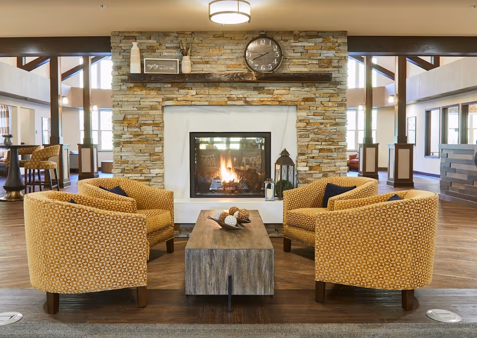A cozy living room area with four patterned yellow armchairs arranged around a wooden coffee table. The background features a stone fireplace with a fire burning inside, a clock above the mantel, and decorative items on the mantel. The room has wooden floors and large windows letting in natural light.
