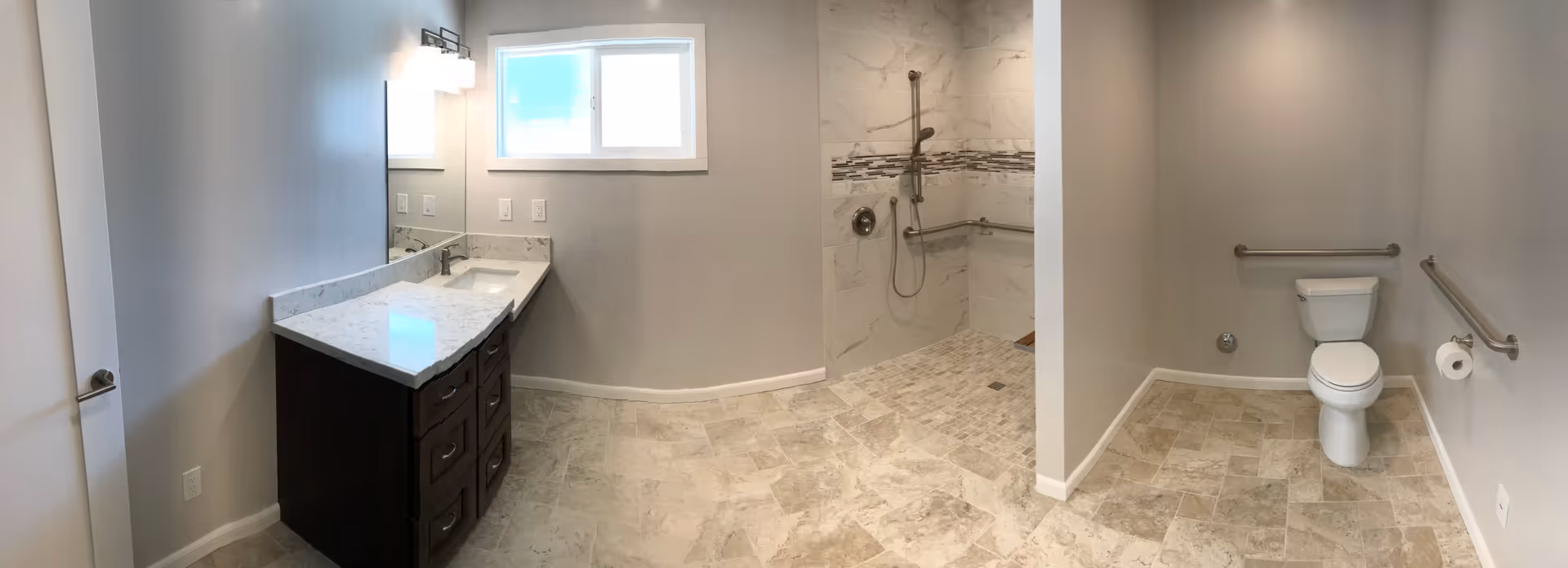A spacious, modern bathroom with beige tiled floor and light gray walls. On the left side, there is a dark wood vanity with a white marble countertop and a rectangular sink beneath a large mirror with two wall-mounted light fixtures. In the center, there is a walk-in shower with a handheld showerhead and grab bars, featuring white and gray marble tiles with a decorative horizontal strip. On the right side, there is a white toilet with grab bars on both adjacent walls and a toilet paper holder.