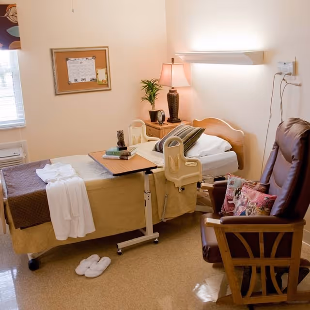 A well-lit senior living bedroom with a hospital-style bed covered in beige and brown bedding, a white robe draped over the bed, and a small overbed table with books and a decorative item. Next to the bed is a wooden nightstand with a lamp, a small plant, and a clock. A comfortable brown recliner chair with colorful pillows is positioned nearby. The room has a window with blinds and a corkboard on the wall.