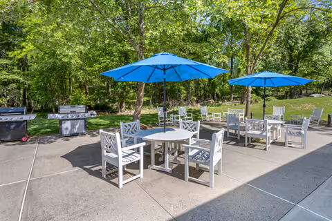 Outdoor patio area with white chairs and tables shaded by blue umbrellas. Two grills are visible on the left side, and green trees and grass surround the area.