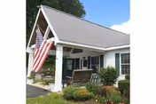 Front entrance of a single-story light-colored building with a covered porch, an American flag, a wooden swing, and landscaped bushes.