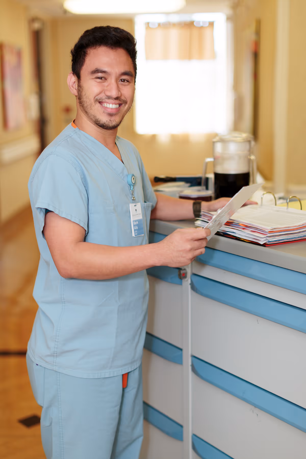 A smiling healthcare worker in light blue scrubs stands at a nursing station or medical cart in a hallway, holding a document and looking at the camera.