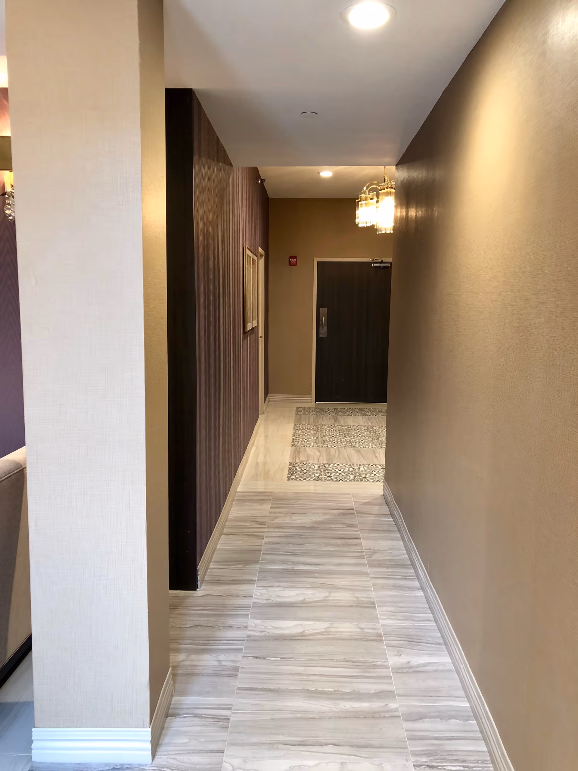 A well-lit hallway with beige walls and light-colored tiled flooring. The hallway leads to a dark wooden door at the end. There are two framed pictures on the left wall and a chandelier-style light fixture hanging from the ceiling near the door.