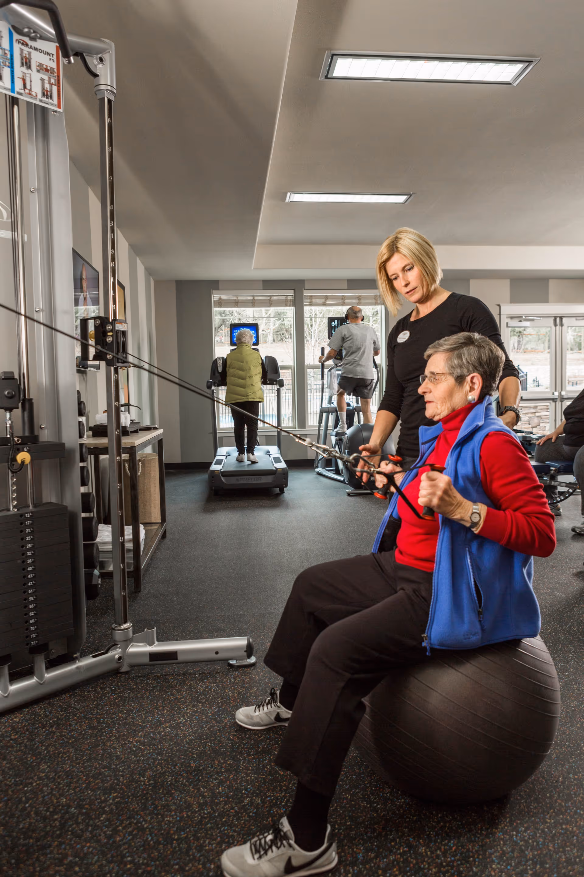 An elderly woman in a red shirt and blue vest is seated on an exercise ball while using a cable machine for strength training, assisted by a female trainer. In the background, two other elderly individuals are using treadmills in a well-lit fitness room with large windows.