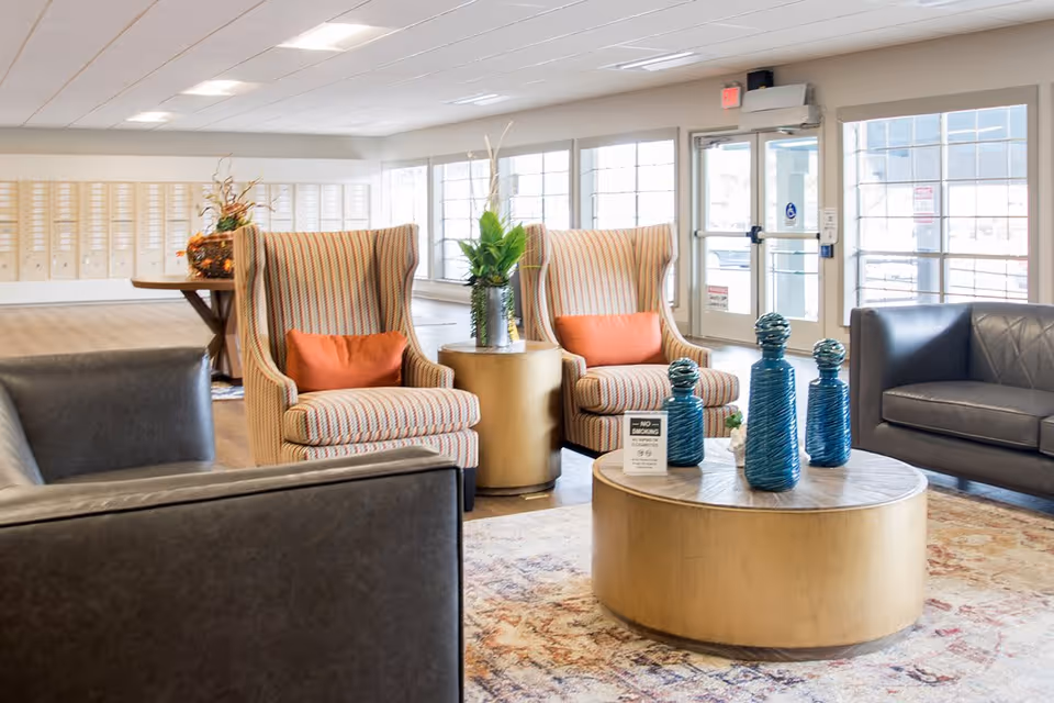 A bright and spacious lounge area with two striped armchairs with orange cushions, two dark leather sofas, a round wooden coffee table with blue decorative vases, and a side table with a green plant. Large windows and glass doors allow natural light to fill the room. Mailboxes are visible along the far wall.