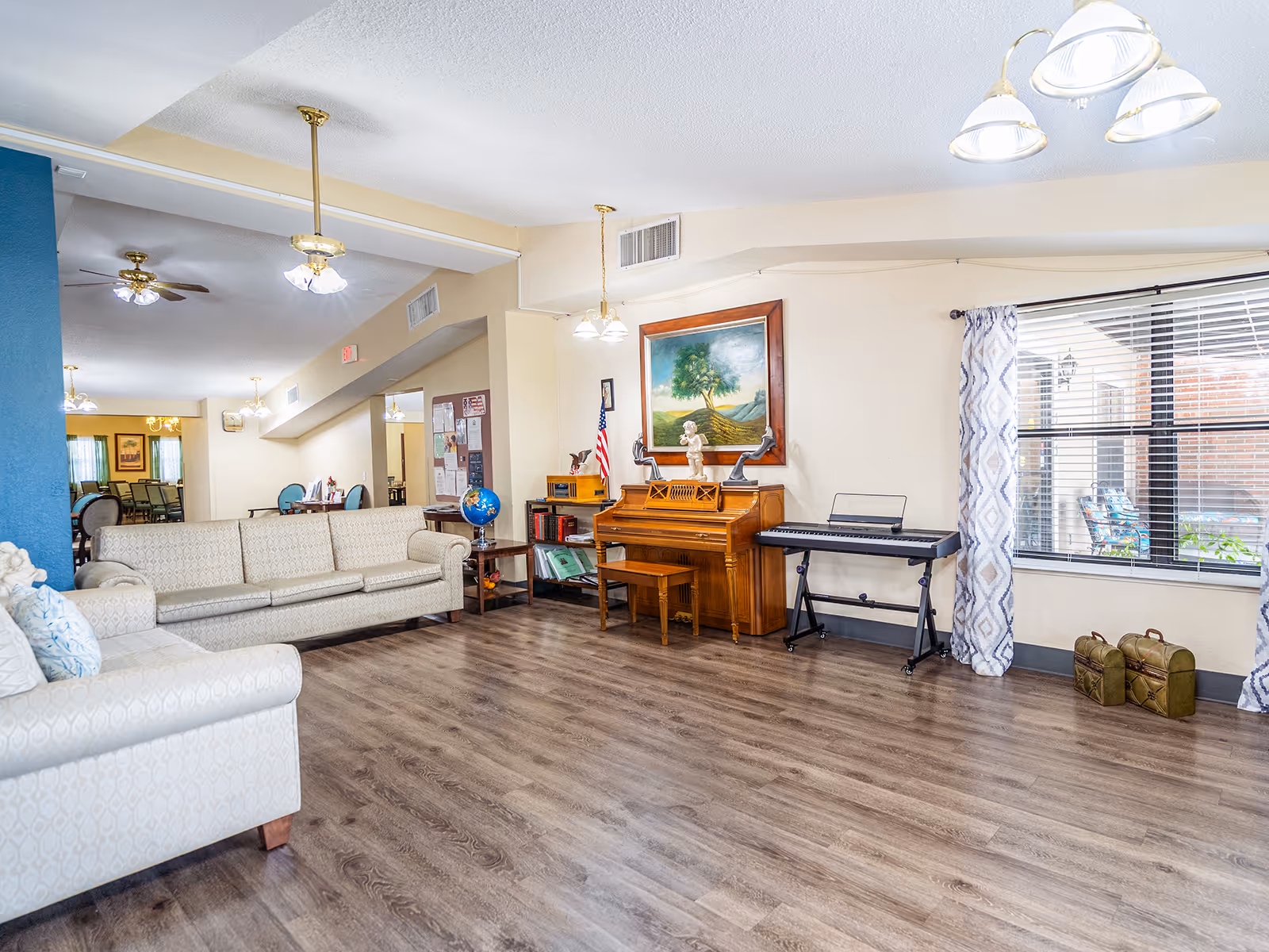 Spacious furnished common living room with sofas, a piano and keyboard, wood-look flooring, and a large window.