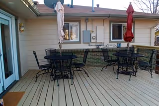 Outdoor patio area with wooden decking, featuring two black metal tables each surrounded by four matching chairs. Each table has a closed umbrella, one beige and one red. The patio is adjacent to a building with beige siding and brick accents, with windows and utility boxes visible on the wall.