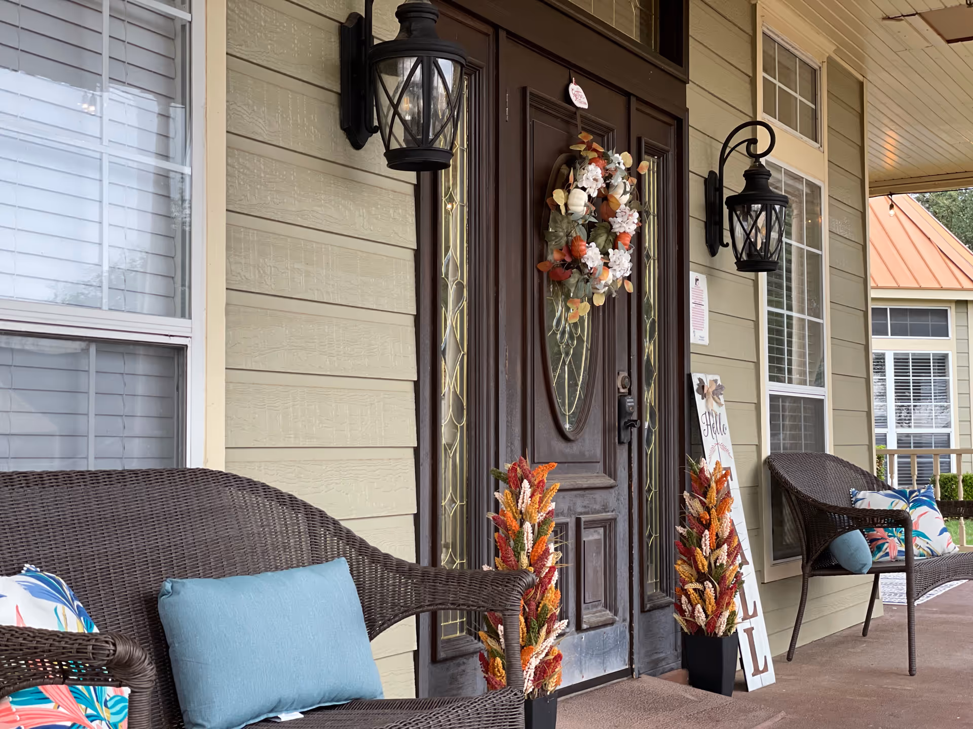 Front porch entrance with a dark wooden door decorated with a floral wreath, flanked by wicker chairs, lantern-style lights, and potted fall decorations.