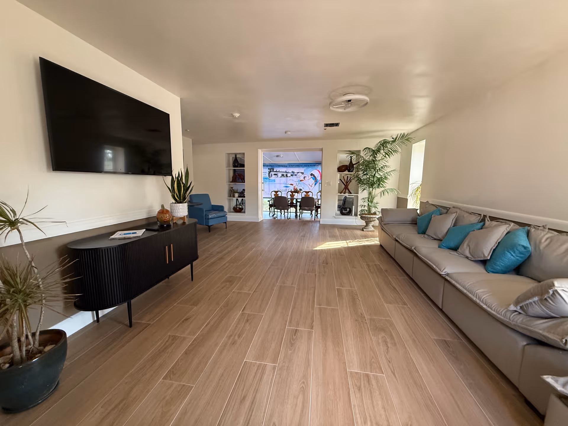 A spacious living room area with light wood flooring, a long gray sofa with multiple blue and gray cushions on the right, a black console table with plants and decorative items on the left, a large flat-screen TV mounted on the wall above the console, and a blue armchair near the back. The room leads to another area with a dining table and chairs, visible through a wide doorway. The walls are white, and there are several plants and decorative shelves in the room.