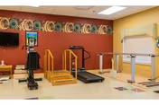 An indoor physical therapy or rehabilitation room with exercise equipment including a treadmill, parallel bars for walking practice, and a stationary exercise bike. The room has a red accent wall with decorative plates displayed near the ceiling and a window with blinds on the adjacent yellow wall.