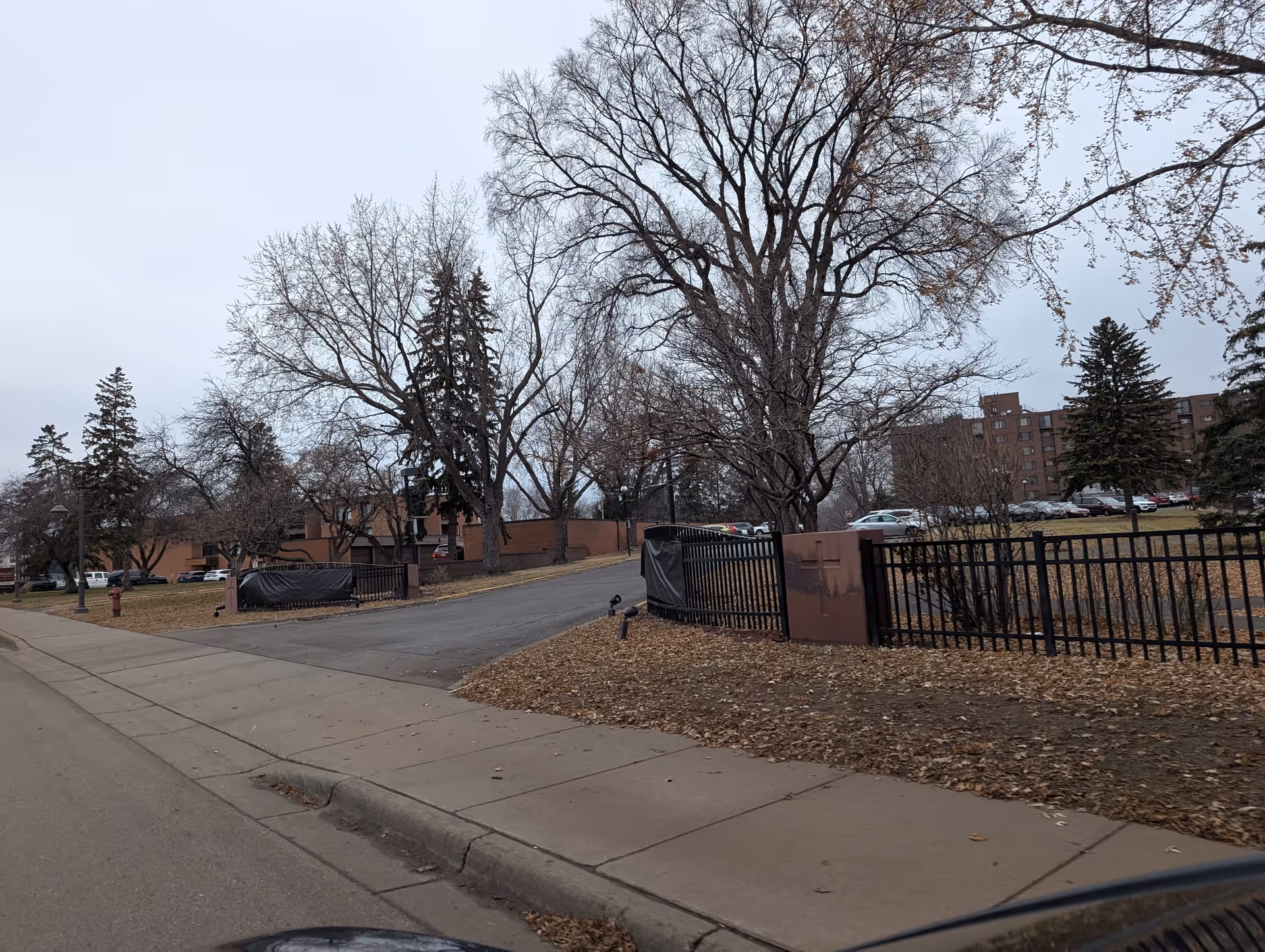 Exterior view of a senior living facility entrance with a metal fence and gate, parking lot and brick buildings behind leafless trees.