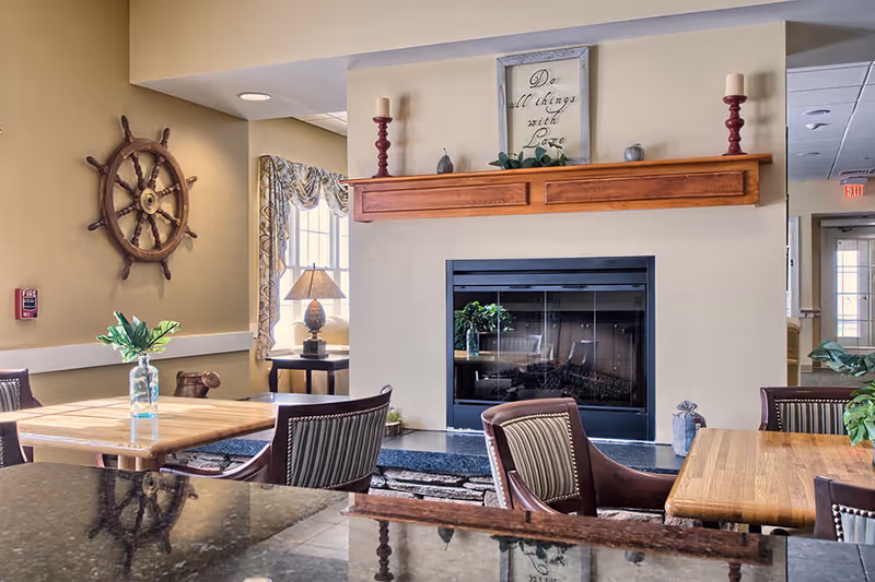 A cozy common area with wooden tables and striped cushioned chairs arranged around a fireplace. Above the fireplace is a wooden mantel decorated with candles, small plants, and a framed sign that reads 'Do all things with Love.' A ship's wheel hangs on the wall to the left, and a window with patterned curtains lets in natural light. A table lamp sits on a small side table near the window.