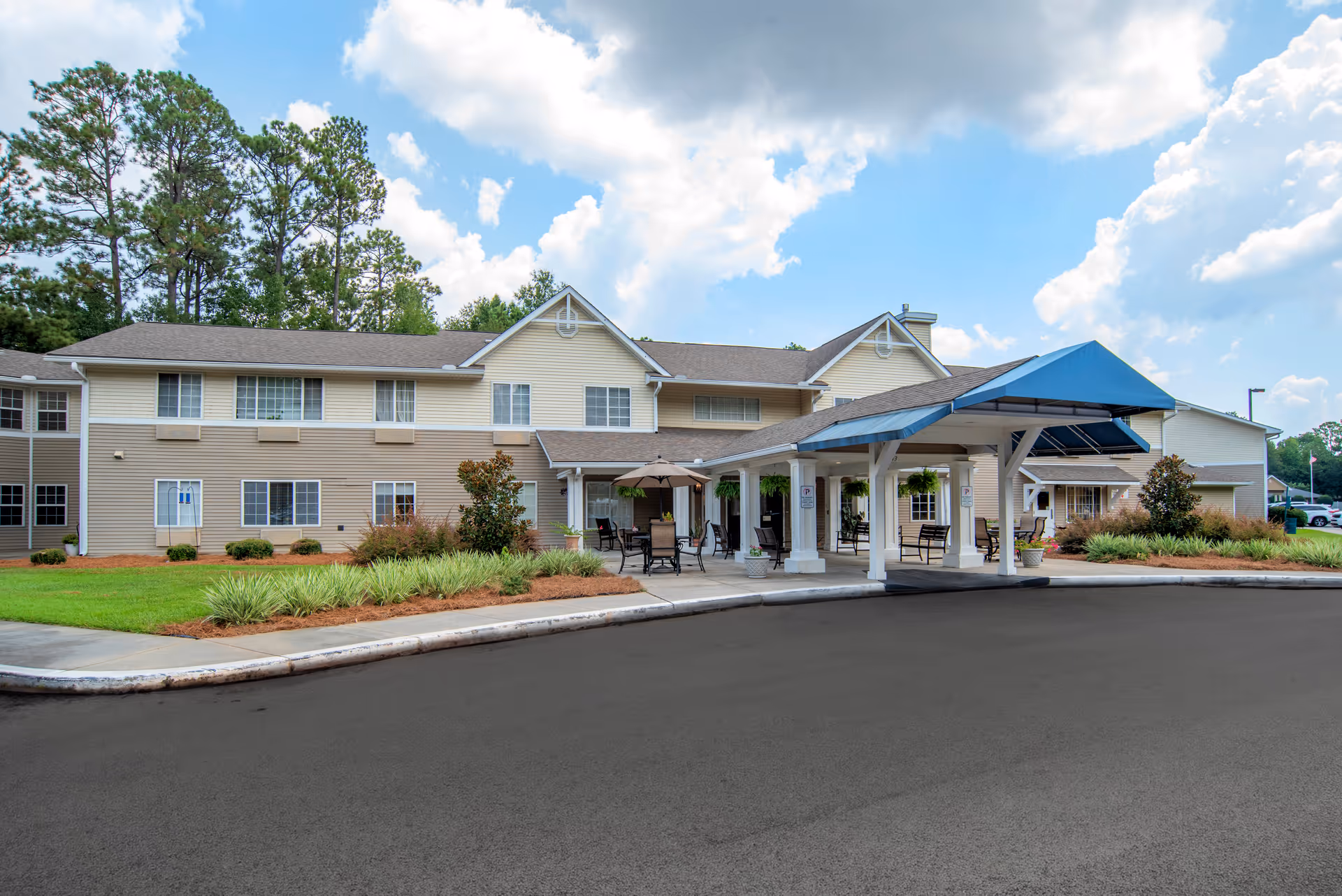 Exterior view of Brookdale Hattiesburg senior living facility showing a two-story building with beige siding, a covered entrance with a blue canopy, outdoor seating with tables and chairs, landscaped greenery, and a partly cloudy sky.