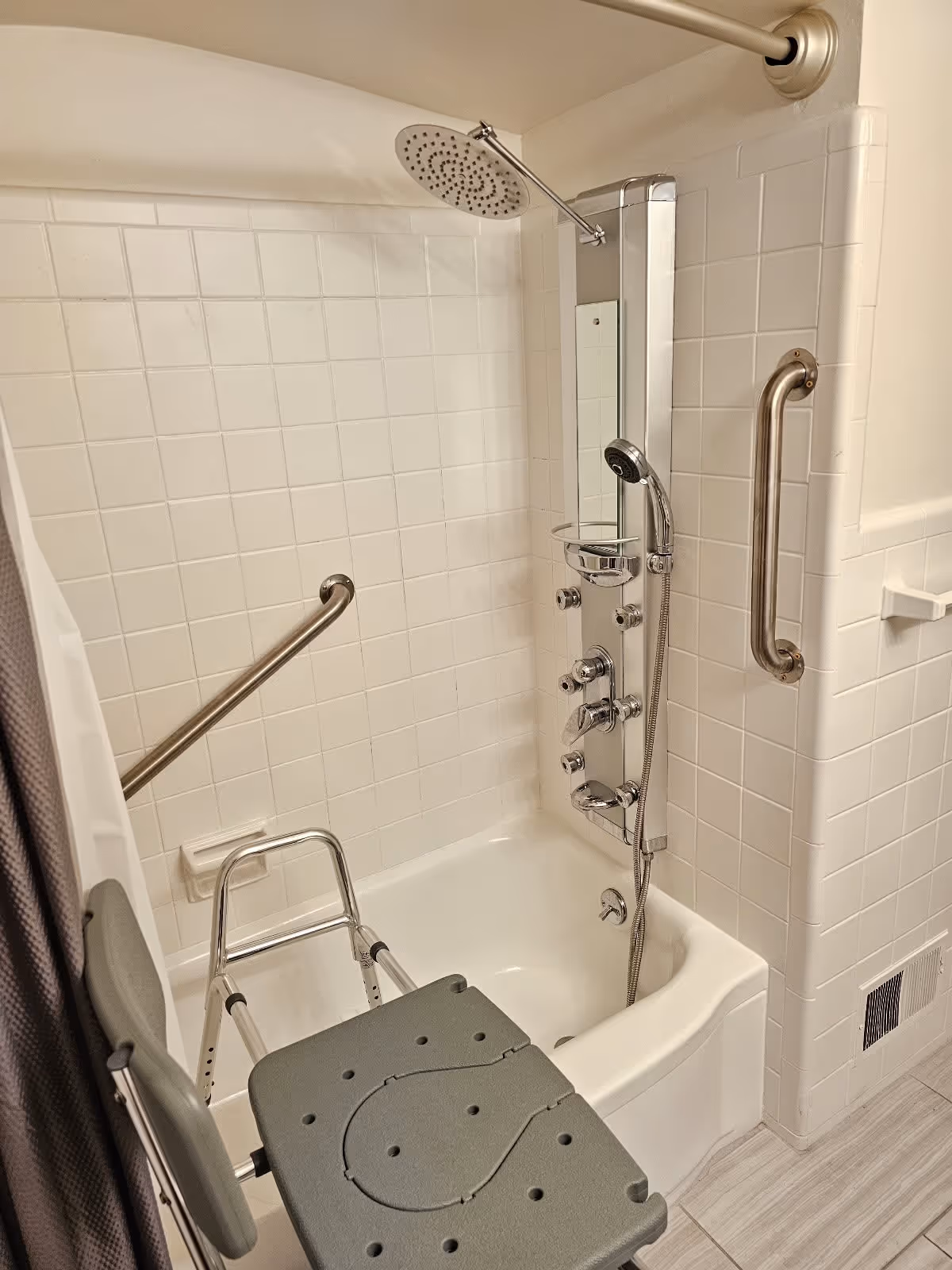 A bathroom shower area with white tiled walls, a bathtub, a mounted shower panel with multiple controls and a handheld showerhead, a large overhead rain showerhead, and two metal grab bars for support. There is also a gray shower chair placed inside the bathtub.