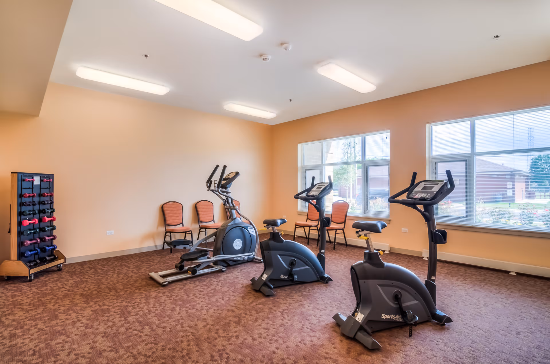 A fitness room with exercise equipment including two stationary bikes and an elliptical machine. There are three chairs against the wall and a rack of colorful dumbbells in the corner. The room has large windows letting in natural light and beige walls with a brown carpeted floor.