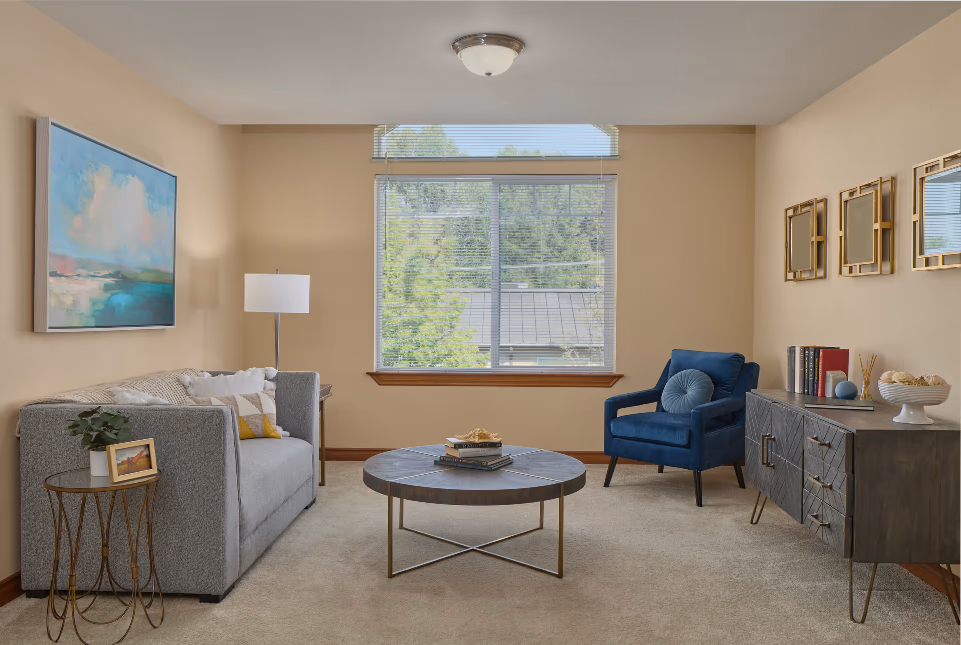 Cozy living room with a gray sofa, blue armchair, round coffee table, large window, and sideboard with decor.