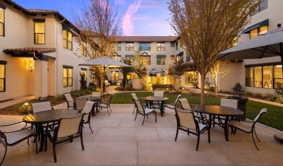 Outdoor courtyard patio with round tables, chairs and umbrellas surrounded by landscaping and a U-shaped three-story building at dusk.