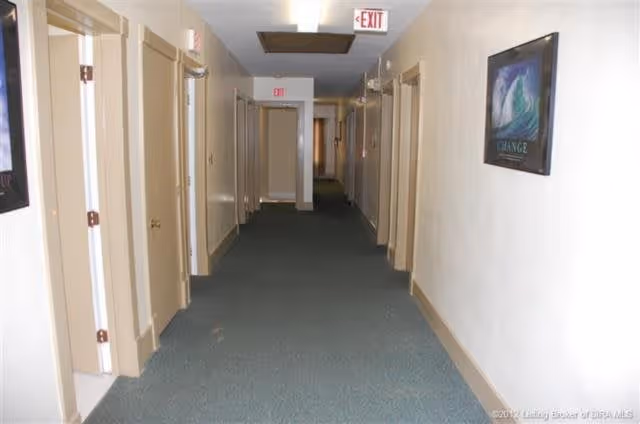 A long indoor hallway with multiple closed beige doors on both sides, green carpet flooring, white walls, and ceiling lights. There are exit signs visible at both ends of the hallway and a framed picture on the right wall.