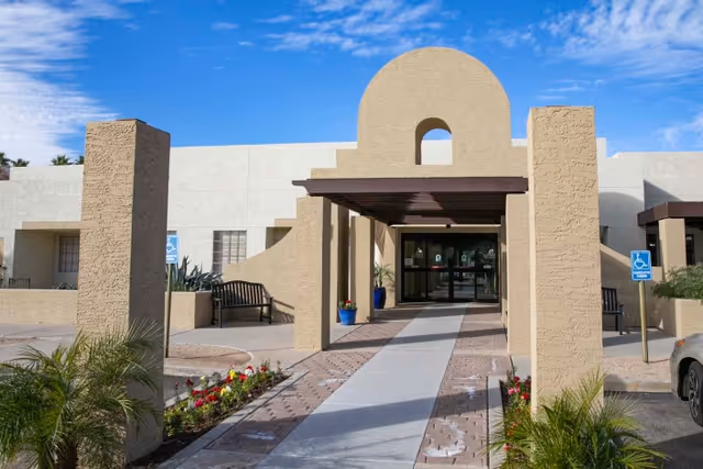 Beige stucco rehabilitation center front entrance with a covered walkway, arched entry, benches, and landscaping.
