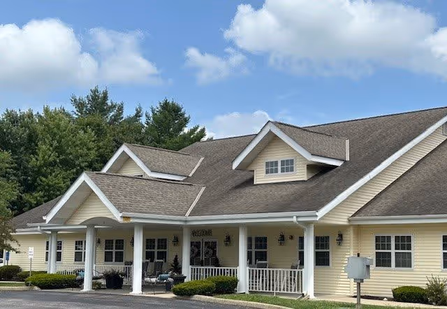 Exterior view of a single-story building with light yellow siding and a dark shingled roof under a partly cloudy sky. The building has a covered entrance with white columns and a small porch area with chairs. There are trees in the background and a paved driveway in front.