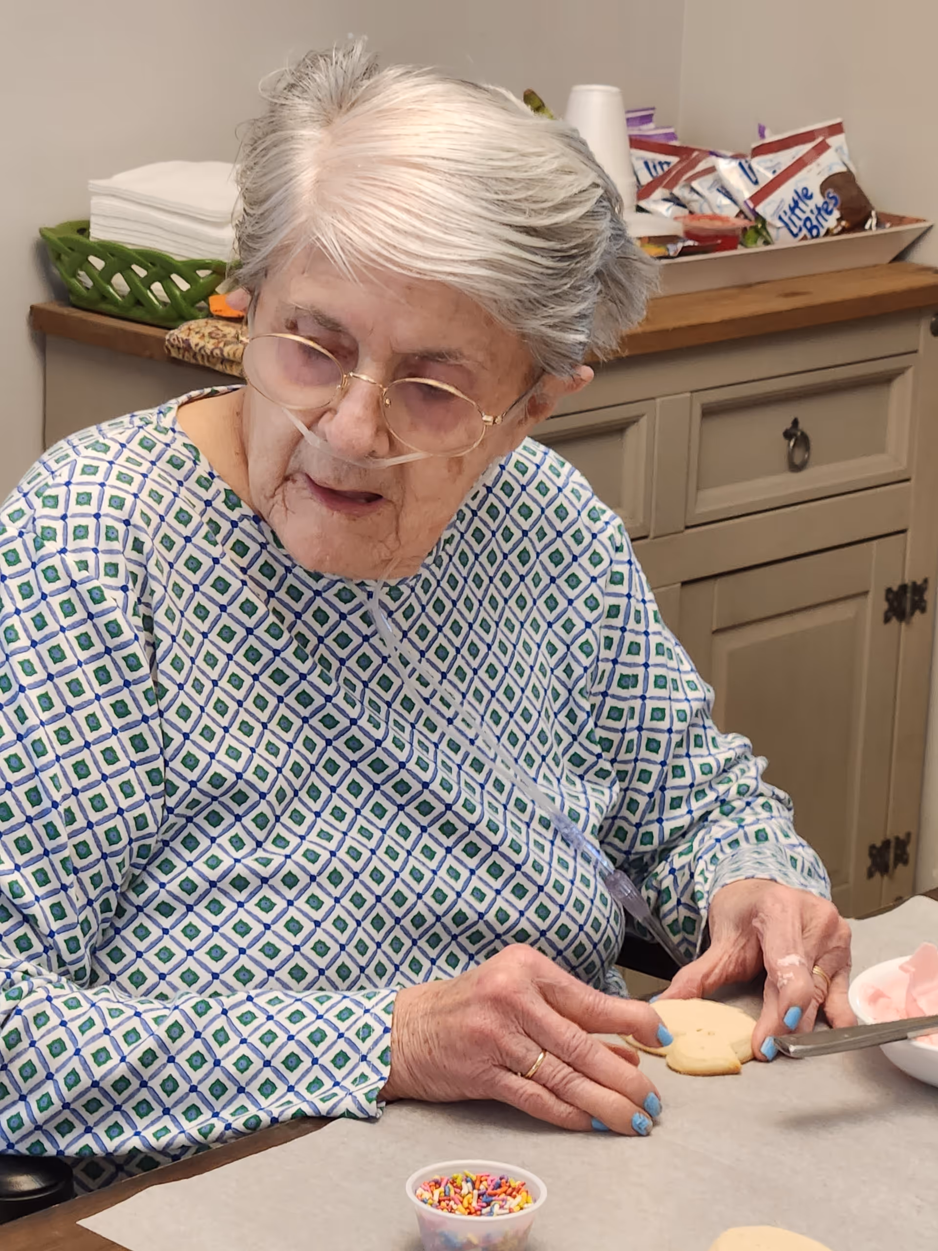 An elderly woman with gray hair and glasses is sitting at a table, decorating a cookie with pink icing. She is wearing a patterned long-sleeve shirt and has a nasal oxygen tube. On the table in front of her are a small container of colorful sprinkles and another cookie. In the background, there is a cabinet with snacks and napkins on top.