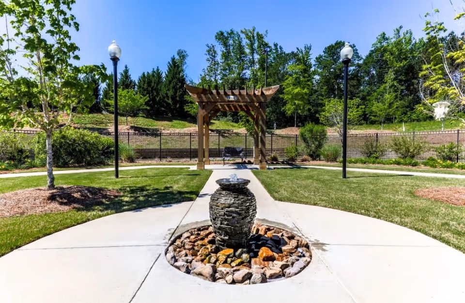 Outdoor garden area with a stone water fountain in the center surrounded by a circular arrangement of rocks. There is a concrete pathway leading to a wooden pergola with a bench underneath. The area is landscaped with grass, trees, bushes, and two lamp posts on either side of the pathway. A black metal fence and dense green trees are visible in the background under a clear blue sky.