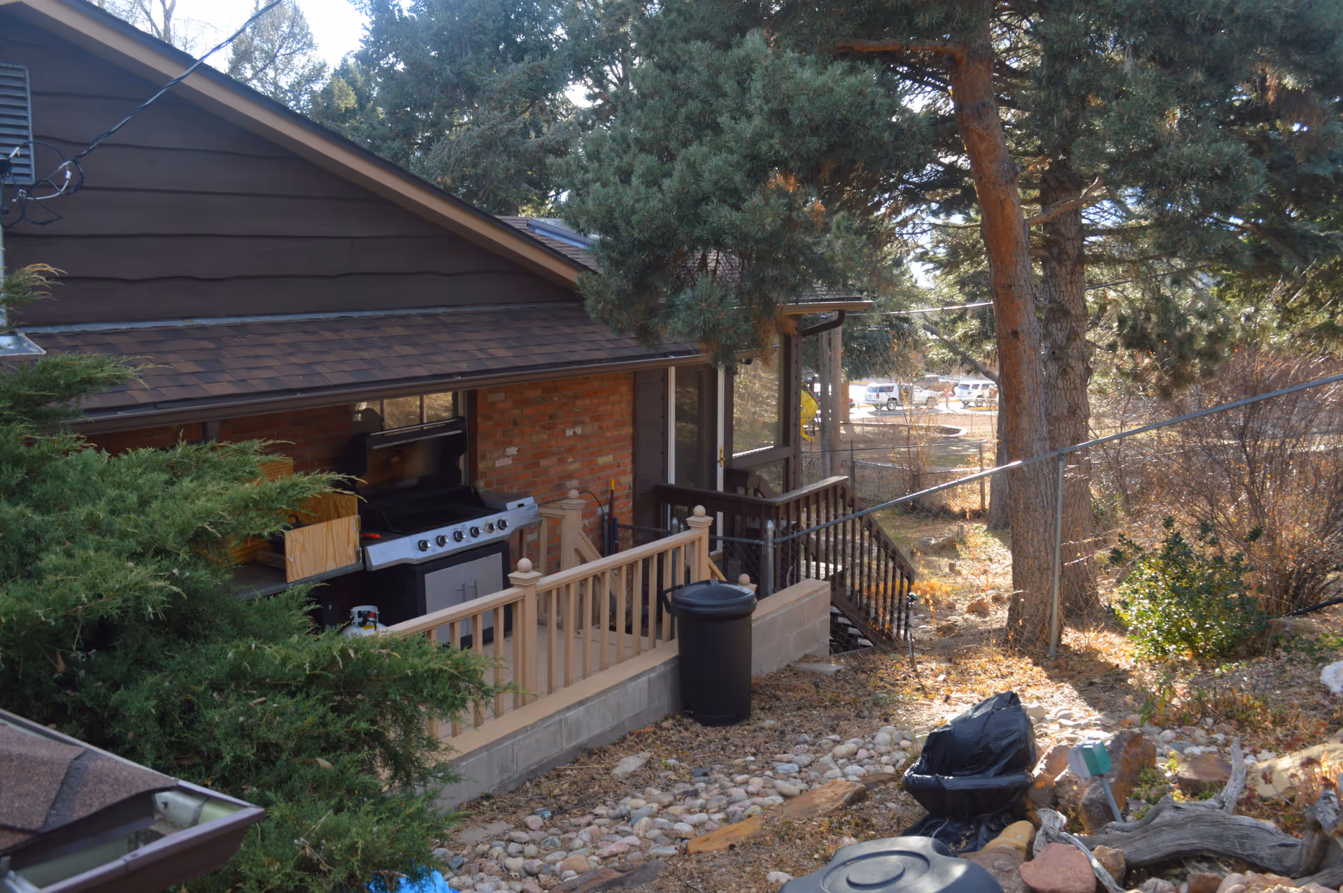 Outdoor view of a residential area with a brown house featuring a brick wall and a porch with a railing. There is a large barbecue grill on the porch, surrounded by trees and bushes. The ground is covered with rocks and dry leaves, and there are some trash bins and a black garbage bag visible. In the background, there are parked cars and more trees.