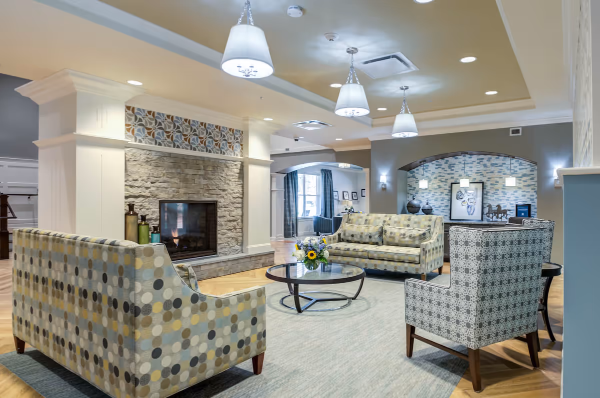 A cozy senior living facility common area with patterned sofas and armchairs arranged around a round glass coffee table with a flower arrangement. The room features a stone fireplace with decorative tiles above it, soft lighting from hanging lamps, and a light blue rug on wooden flooring. The background shows an archway leading to another seating area with artwork and pendant lights.