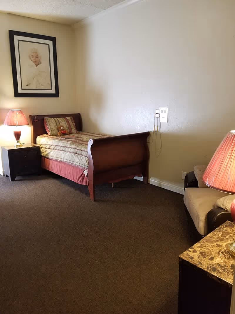 A small bedroom with a single wooden bed covered with a striped bedspread and a pillow. A teddy bear is placed on the bed. There is a nightstand with a red lamp beside the bed. On the right side, there is a cushioned armchair with another red lamp on a marble-topped table. A framed black and white portrait hangs on the wall above the bed. The room has beige walls and brown carpeted floor.